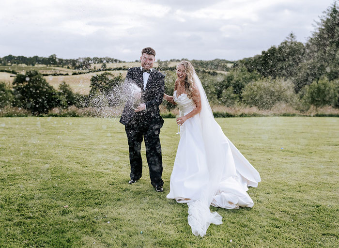 a groom spraying a bottle of champagne as bride laughs. They are standing on grass and there are lots of trees and a field in the background