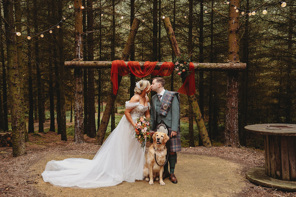 Bride and groom kissing beneath a rustic woodland wedding arch with their golden retriever in Scotland