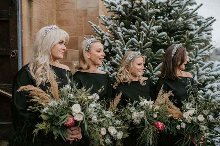 four women wearing black dresses are holding bouquets of white and pink flowers while standing in front of a blonde sandstone building and a snowy fir tree