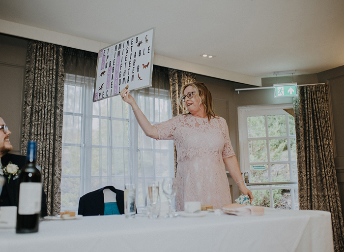 A woman in a pink dress holds up a sign to people sitting at the table in front of her