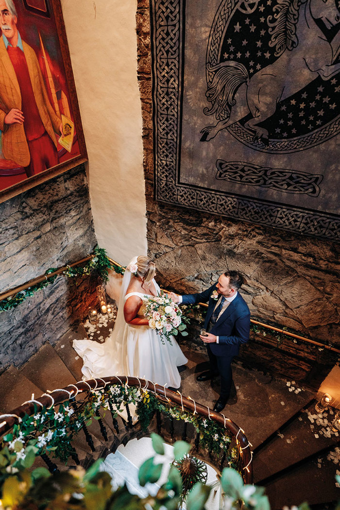 overhead view of bride and groom stood facing each other on a curved stone staircase with twinkling lights wrapped around the bannister 
