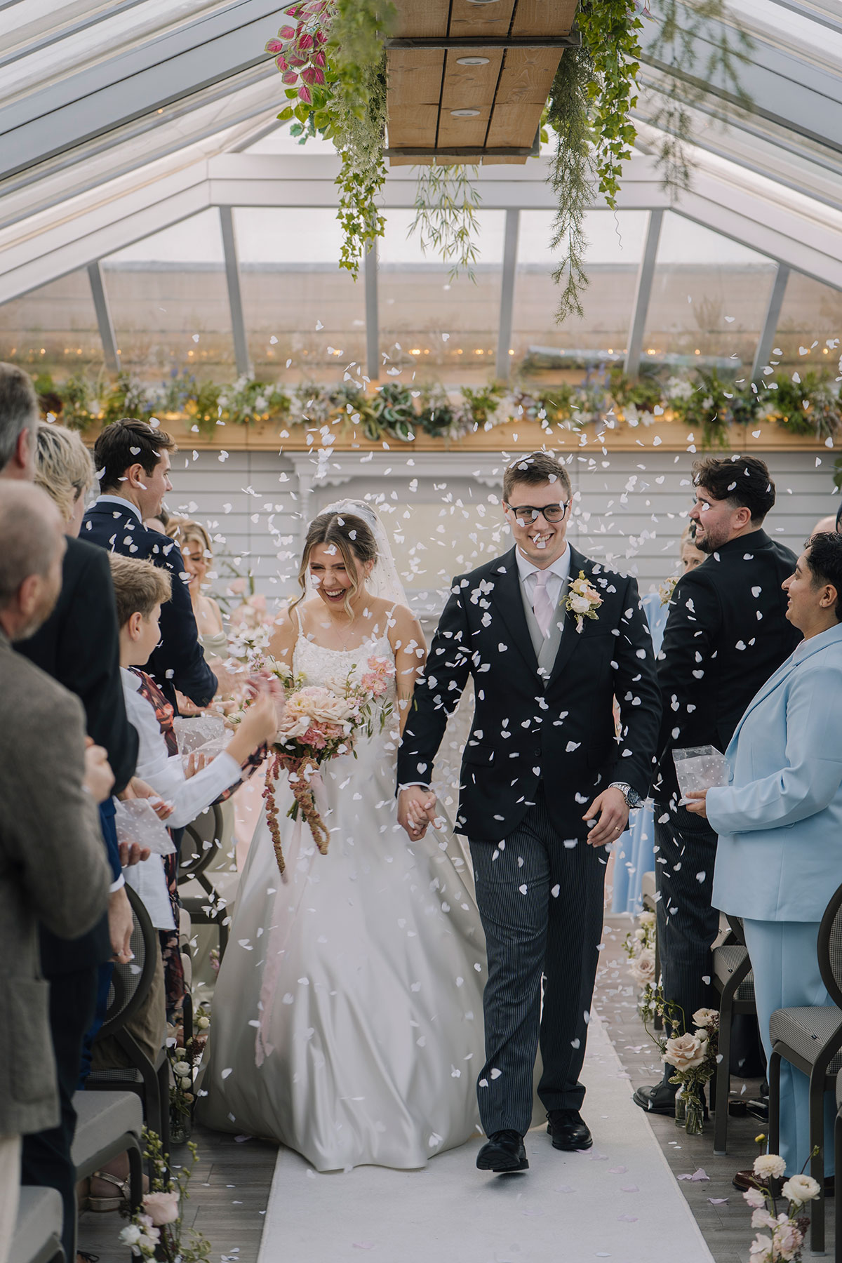 Newlywed couple walking down the aisle as guests throw confetti during a joyful indoor ceremony exit
