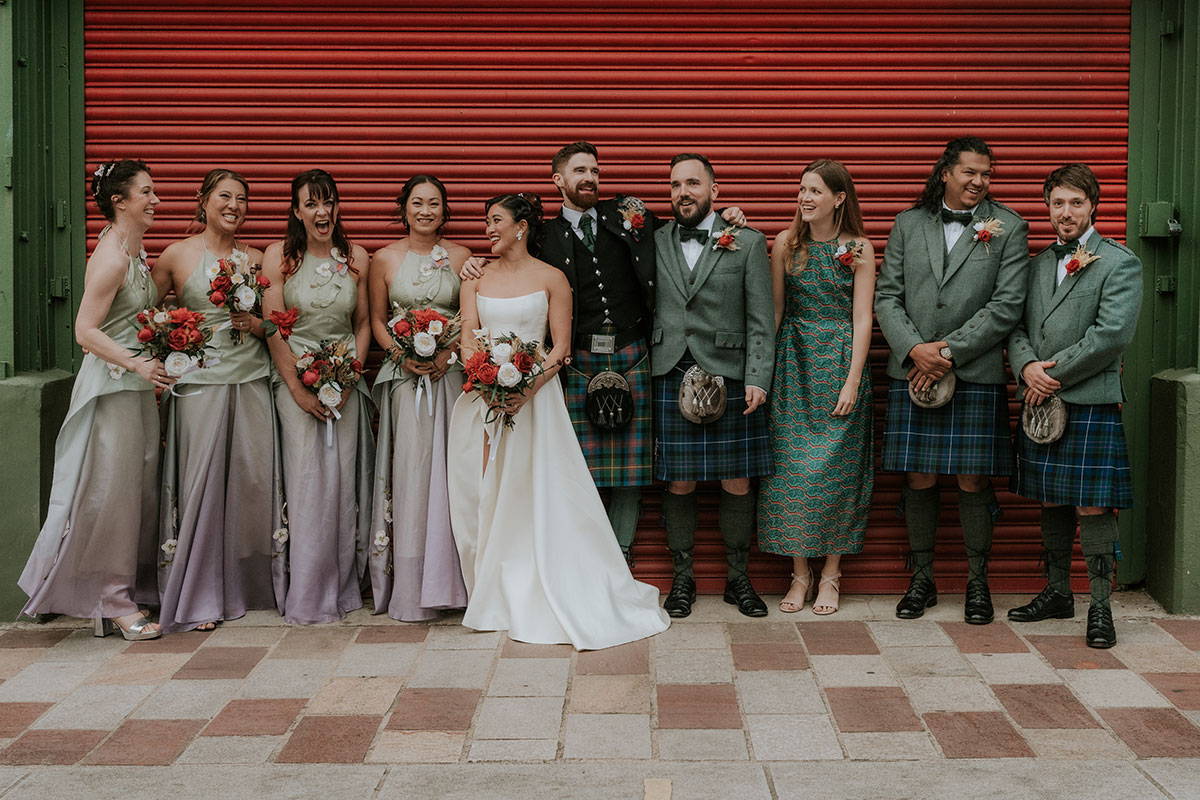 Bride and groom with bridal party lined up against red shutter at BAaD Glasgow in coordinated outfits