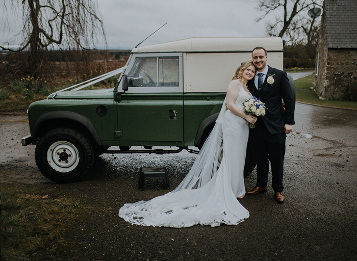 A bride in a fitted dress and long veil and groom in a suit stand in front of a green jeep