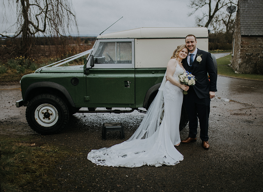 A bride in a fitted dress and long veil and groom in a suit stand in front of a green jeep