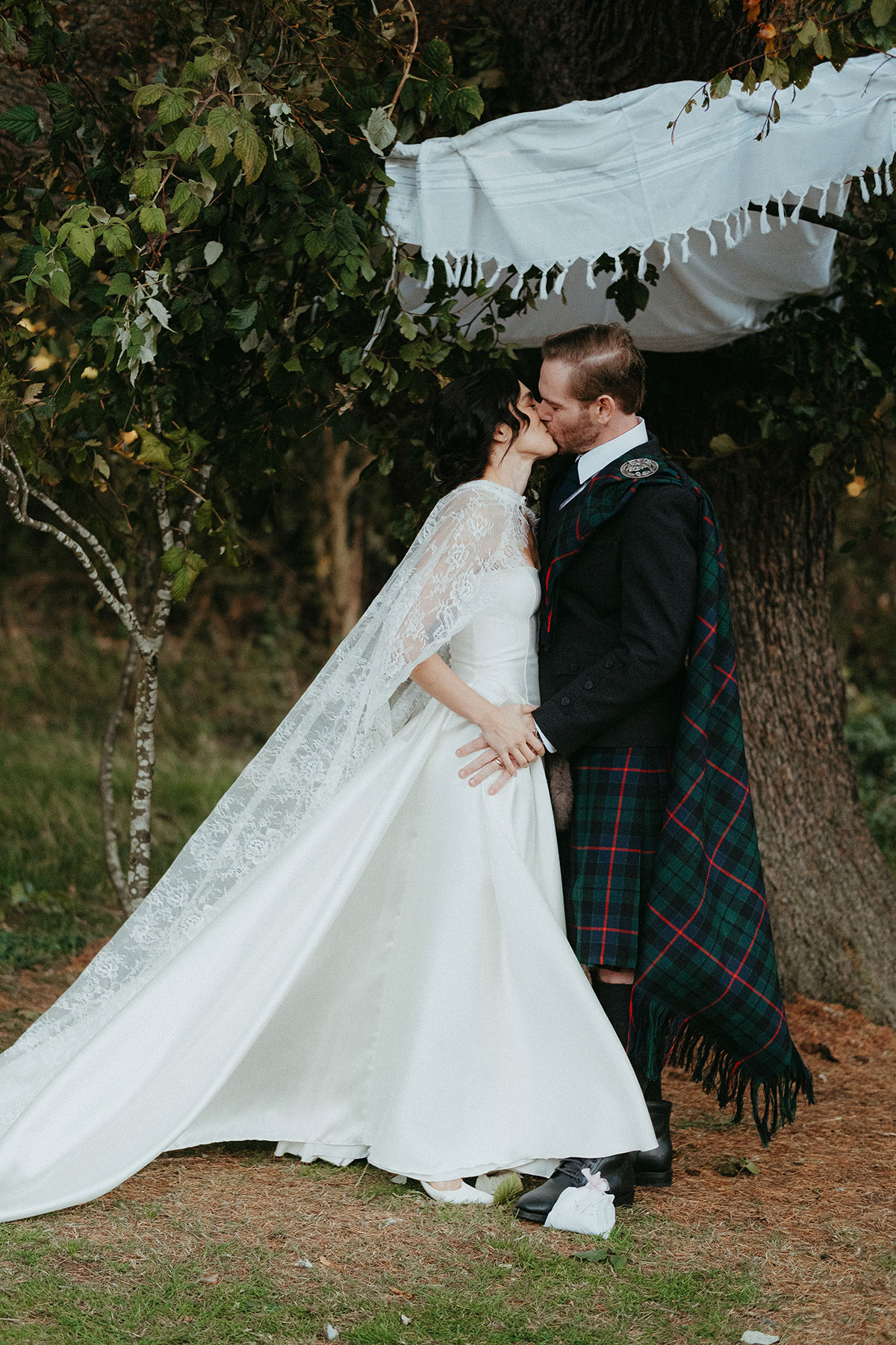 Newlyweds share a kiss beneath a floral chuppah during their outdoor wedding ceremony at Cambo Country House and Estate.