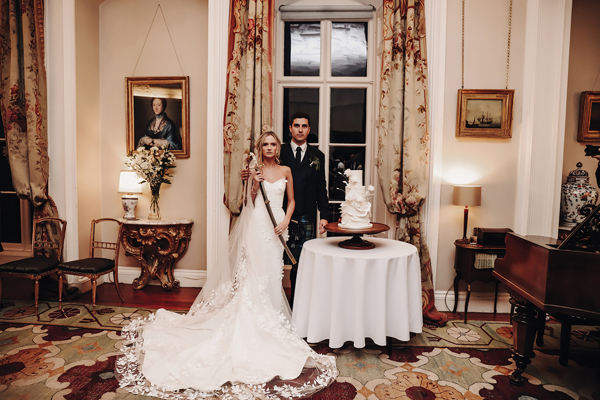 A Bride Holding A Sword Poses With A Groom Next To A Wedding Cake at Winton Castle