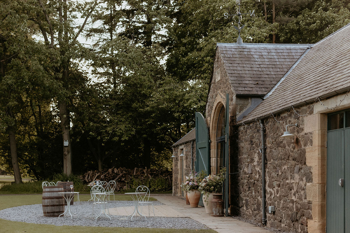 The exterior of a stone barn with open green doors, outdoor seating and surrounding woodland.