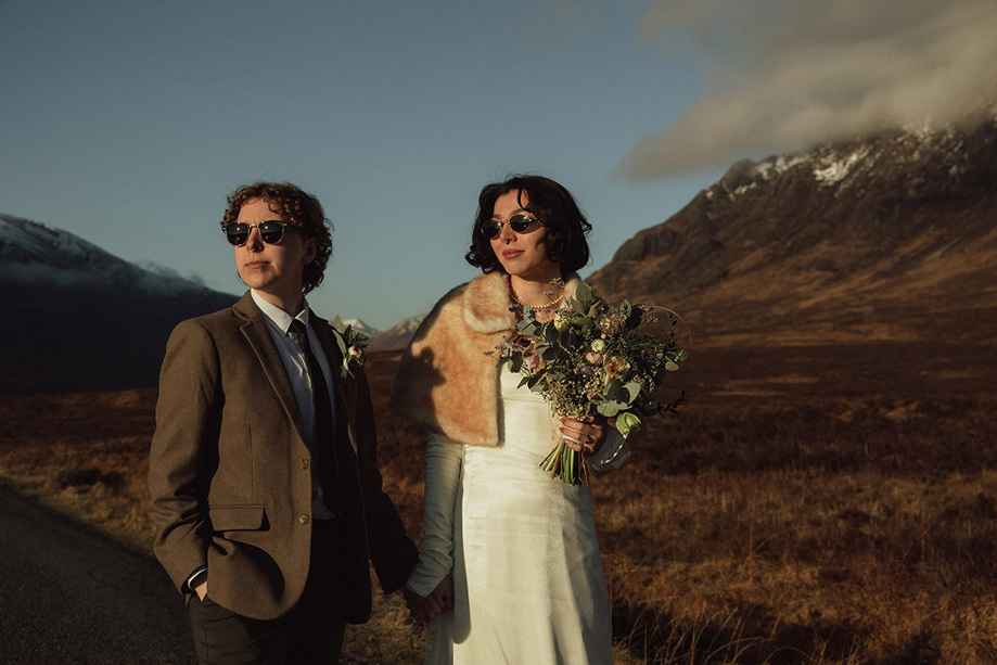 Two brides wearing sunglasses and standing amongst hills with wildflower bouquet