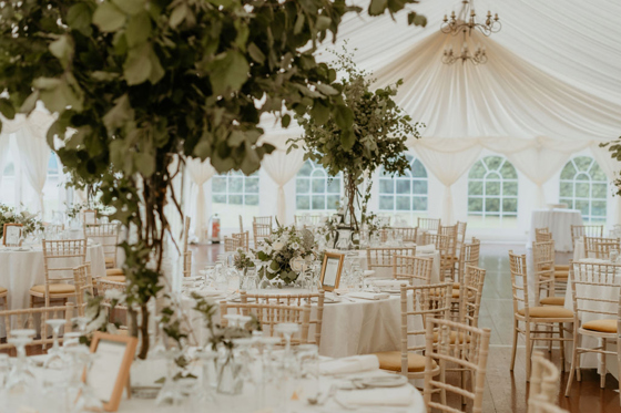 Marquee wedding reception table setup with white linens, chiavari chairs and overhead greenery installations