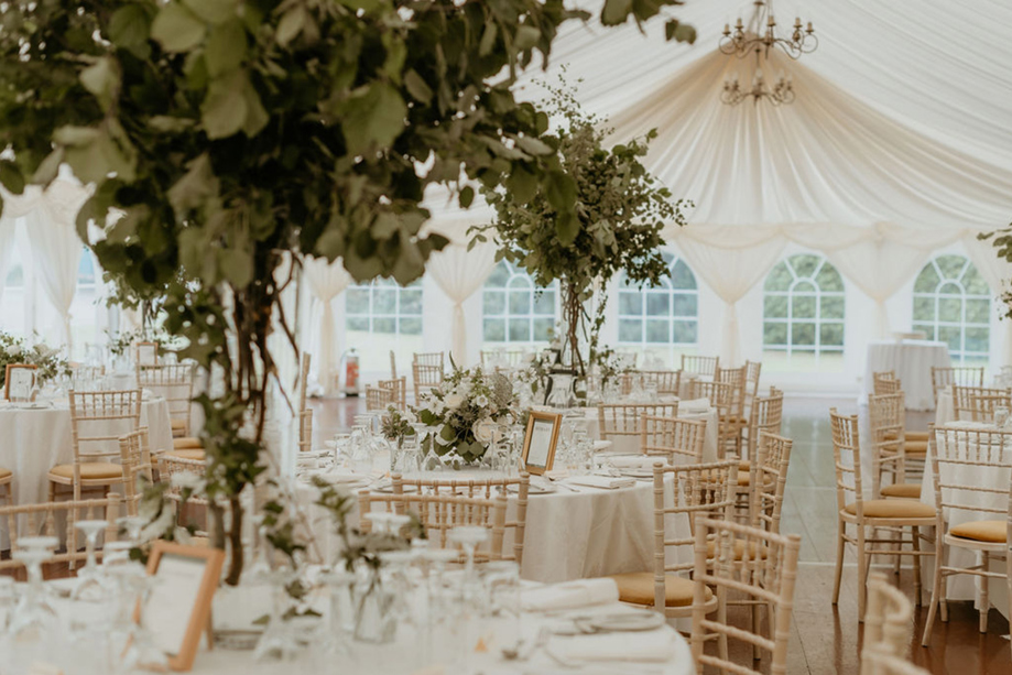 Marquee wedding reception table setup with white linens, chiavari chairs and overhead greenery installations
