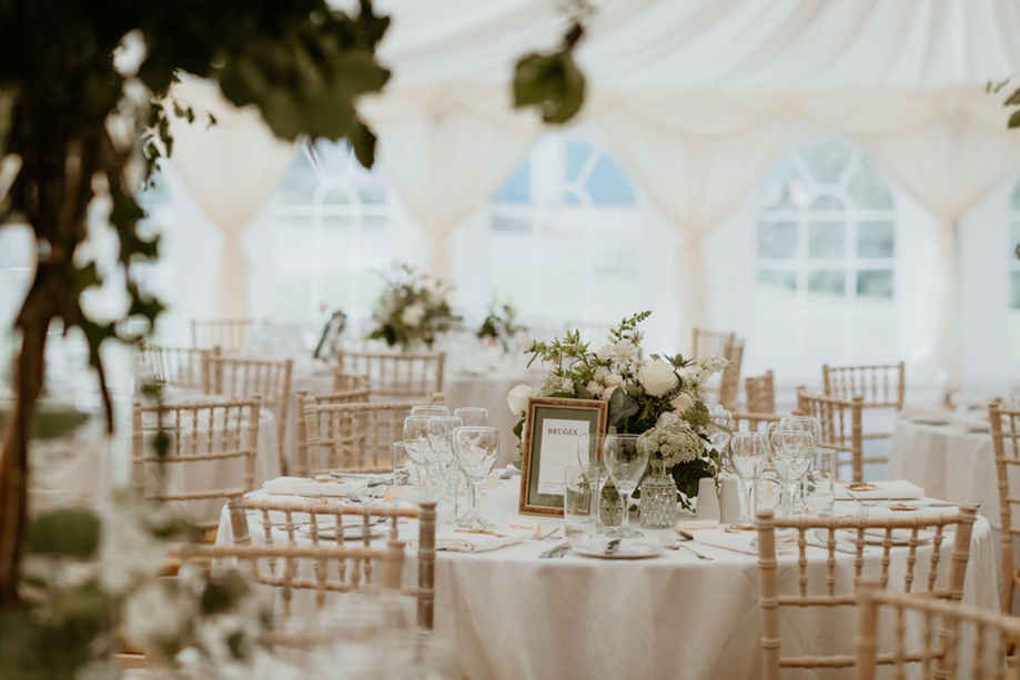 Wedding breakfast tables styled with white rose centrepieces and chiavari chairs inside a light-filled marquee