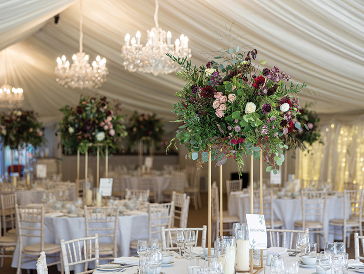 Statement floral centrepiece on gold stand styled on wedding reception tables inside a marquee