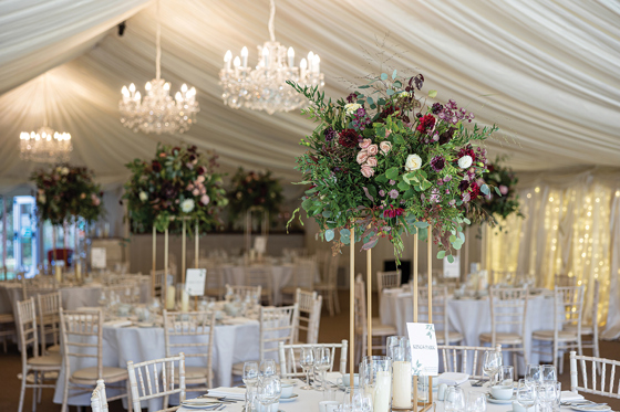 Statement floral centrepiece on gold stand styled on wedding reception tables inside a marquee