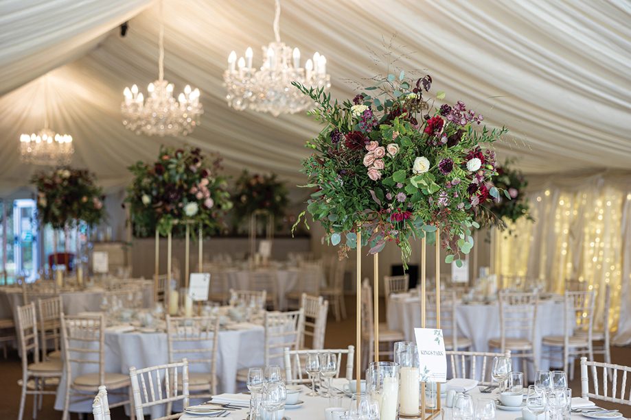 Statement floral centrepiece on gold stand styled on wedding reception tables inside a marquee