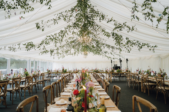 Long marquee wedding table setup with wooden chairs, seasonal florals and hanging greenery overhead