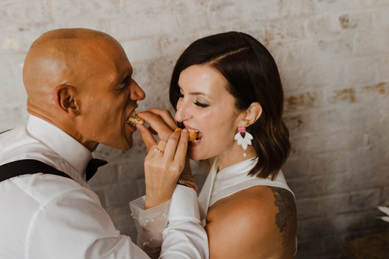 Bride and groom eat macarons, bride is wearing earrings with pink love heart post and pearlescent earring with pearl hanging at bottom