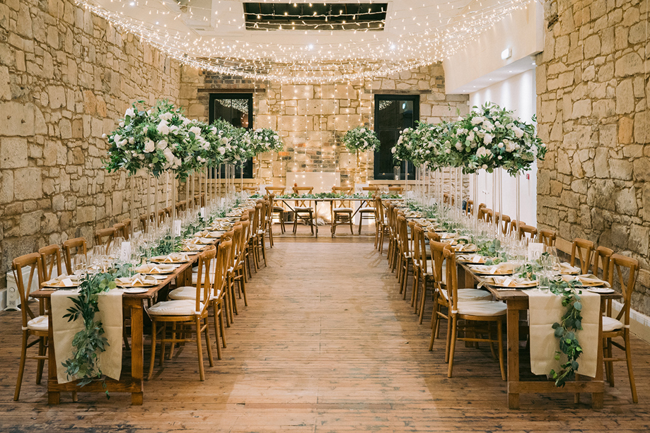 Stone-walled wedding reception with long wooden tables, white flowers and fairy lights planned by Pebblefish Occasions & Events.