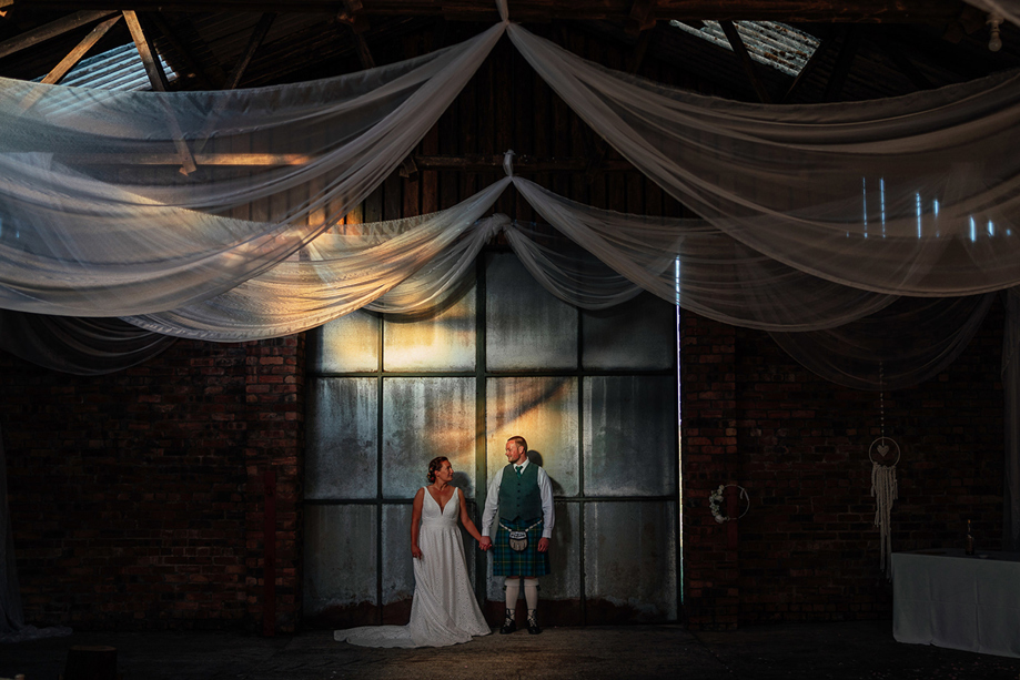 Bride and groom holding hands beneath draped fabric in an industrial wedding venue planned by Pebblefish Occasions & Events.