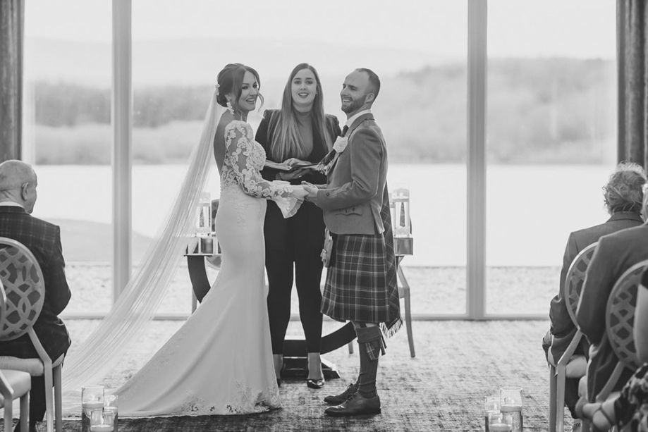 Black and white image of smiling couple holding hands at altar with celebrant