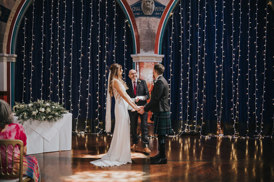 Bride, groom and celebrant smile at altar in front of fairy light curtain and arches