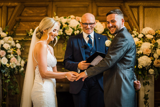 Smiles from bride, groom and celebrant as bride puts grooms wedding ring on