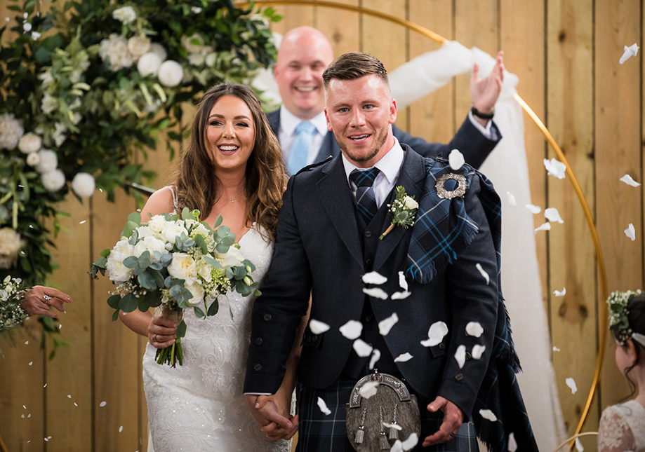 Just married Bride and Groom walk back down the aisle through confetti with smiling celebrant in background