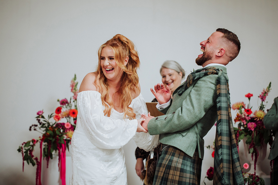 Bride and groom laughing during a joyful wedding ceremony as they hold hands at the altar, surrounded by floral arrangements.
