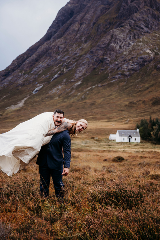Groom carrying his bride over his shoulder during a scenic outdoor elopement in the Scottish Highlands with mountains in the background.