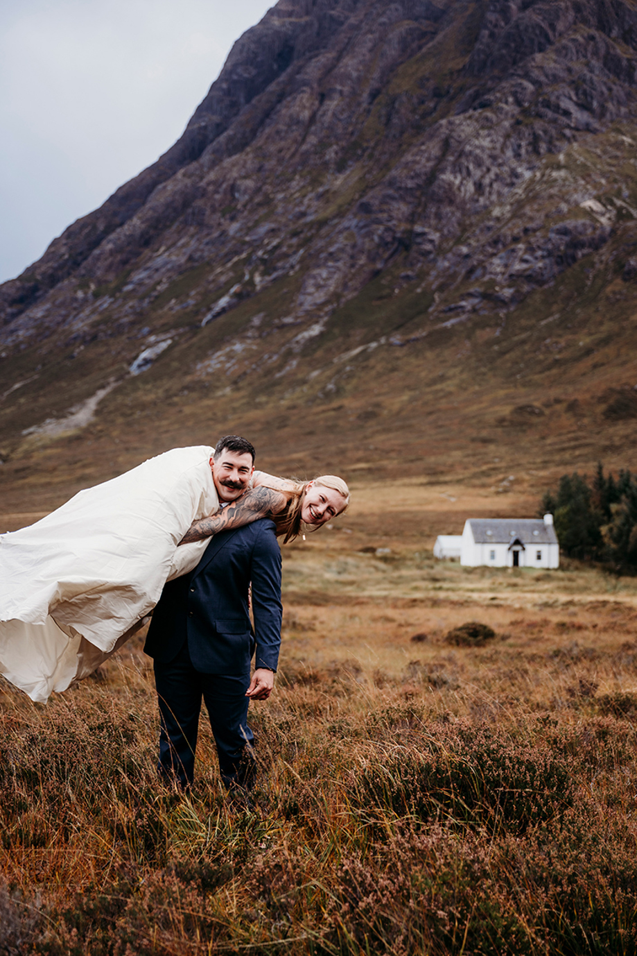 Groom carrying his bride over his shoulder during a scenic outdoor elopement in the Scottish Highlands with mountains in the background.