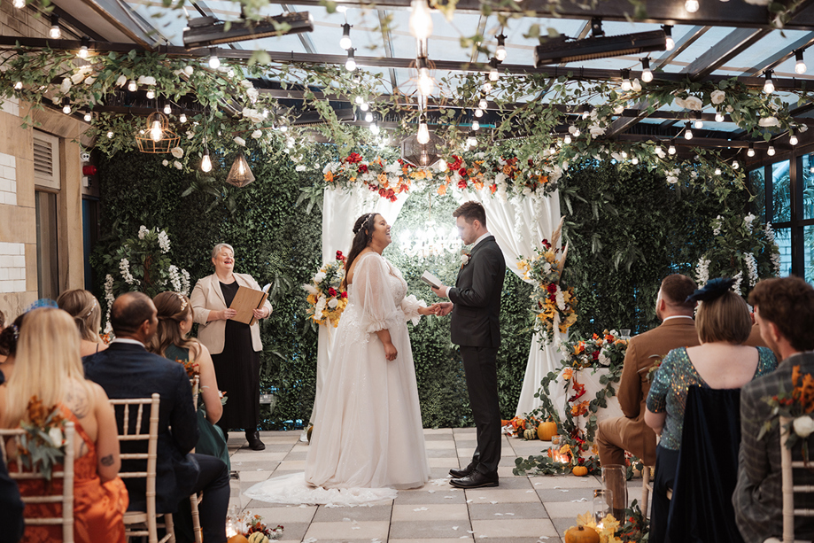 Bride and groom exchanging vows during an intimate wedding ceremony under a floral arch with guests seated nearby.