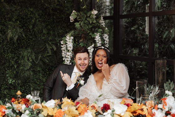 Playful bride and groom seated at the wedding reception table, showing their wedding rings and celebrating with floral décor.