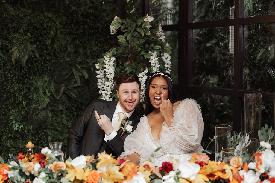 Playful bride and groom seated at the wedding reception table, showing their wedding rings and celebrating with floral décor.