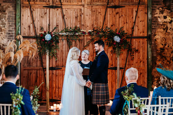 Scottish barn wedding ceremony at Pratis Barns, with a humanist celebrant officiating as the couple exchange vows beneath a floral arch during an indoor rustic wedding.