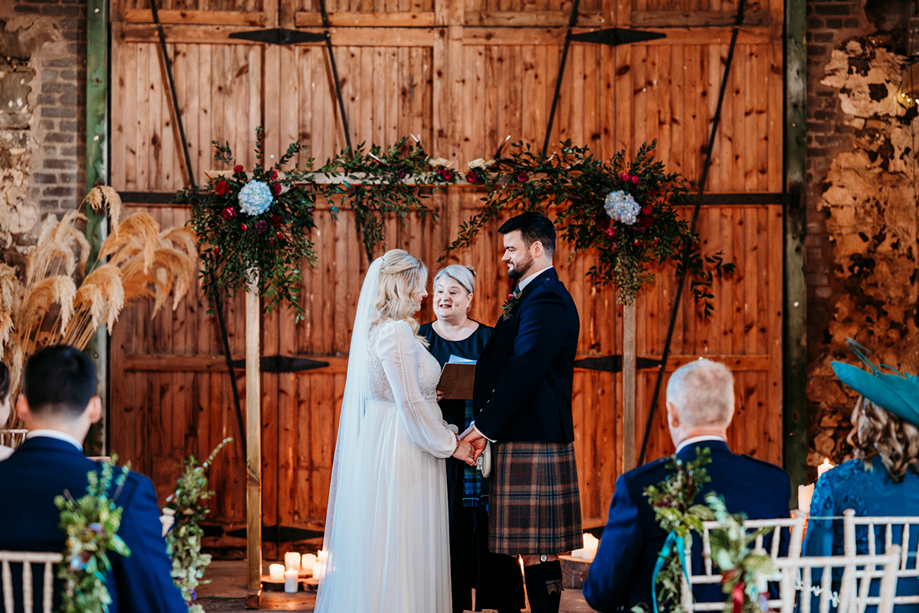 Scottish barn wedding ceremony at Pratis Barns, with a humanist celebrant officiating as the couple exchange vows beneath a floral arch during an indoor rustic wedding.