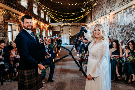 Traditional Scottish handfasting ceremony inside a stone barn, as the bride and groom tie the knot with tartan fabric during a humanist wedding celebration.