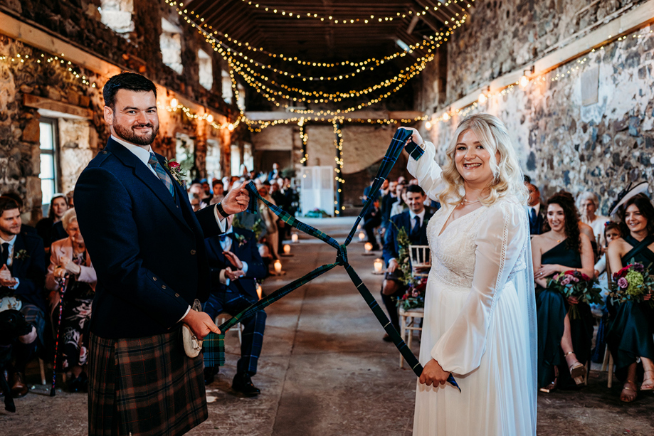 Traditional Scottish handfasting ceremony inside a stone barn, as the bride and groom tie the knot with tartan fabric during a humanist wedding celebration.