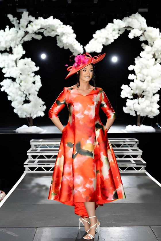 Model in orange floral occasion dress and statement hat on wedding show catwalk