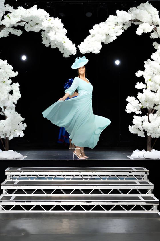 Model in pale teal occasion dress and hat on wedding show catwalk runway