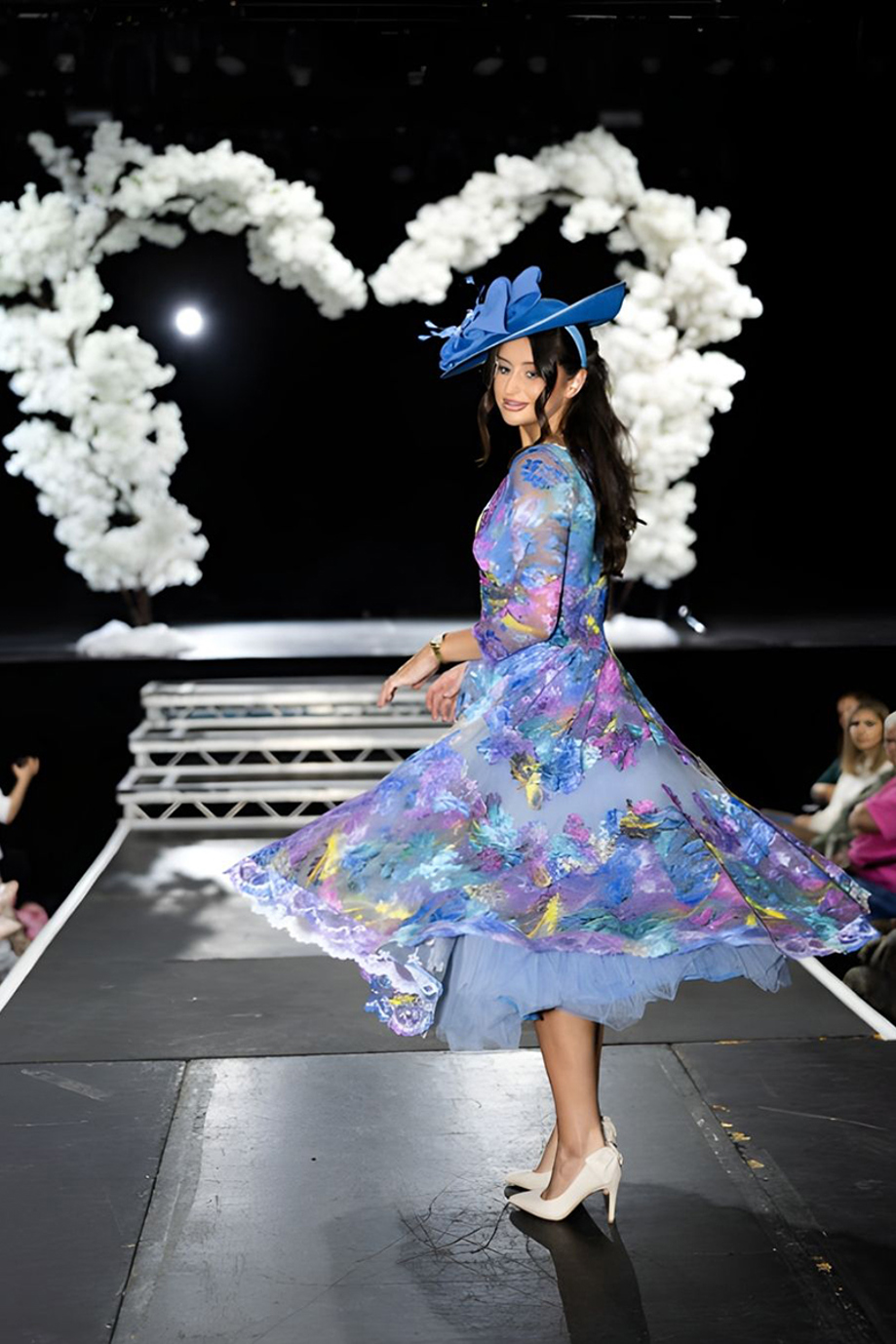 Model twirling in blue and pastel floral midi dress with matching hat on wedding show catwalk