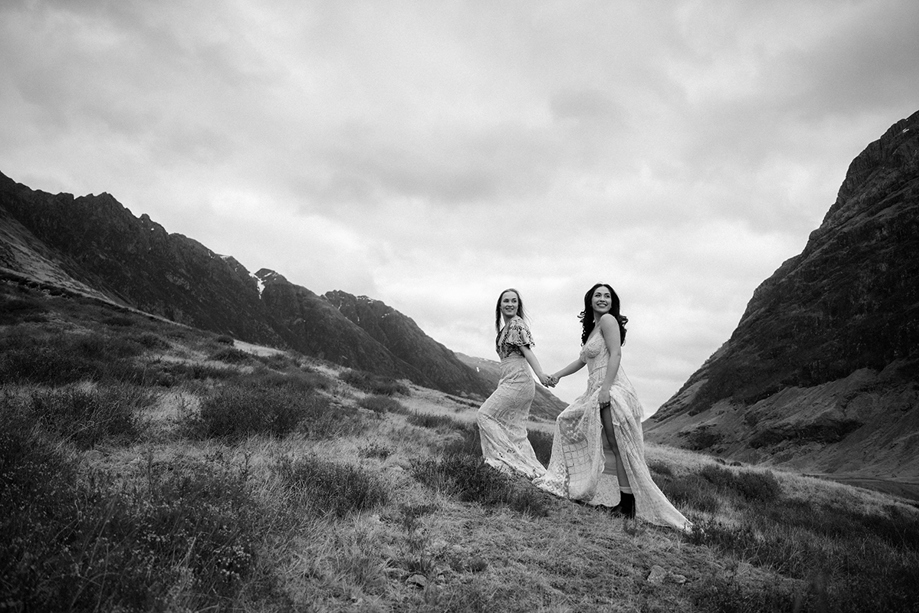 Black and white image of same-sex couple walking hand in hand through Glencoe landscape on elopement-style wedding shoot