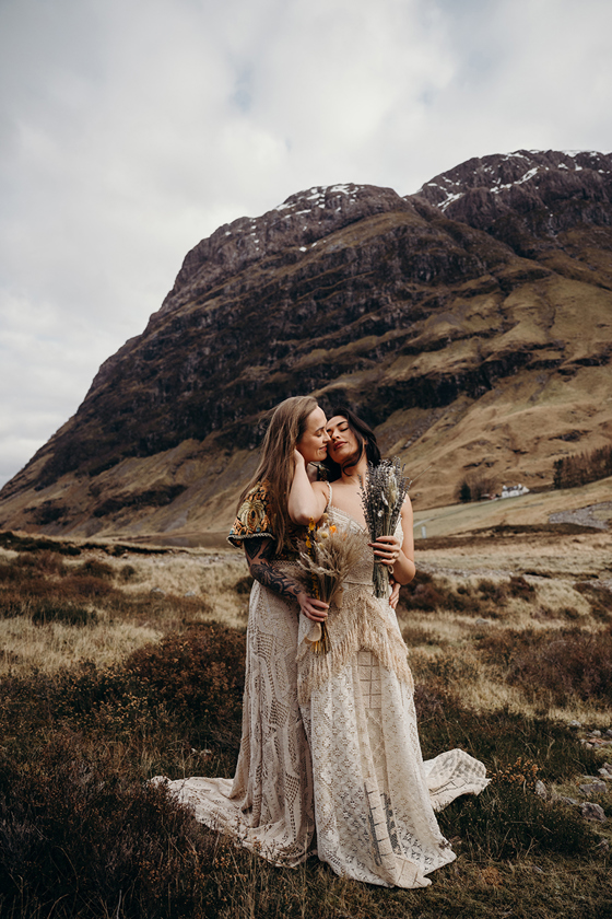 Same-sex couple in boho wedding dresses embracing in Glencoe with dramatic mountain backdrop in the Scottish Highlands