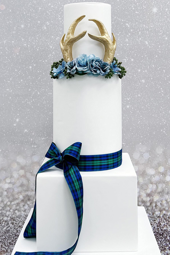 White three-tier cake with blue and green tartan bow, flowers and gold antlers