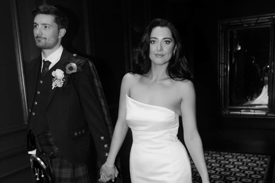 Black and white image of bride and groom walk holding hands with bride smiling at camera