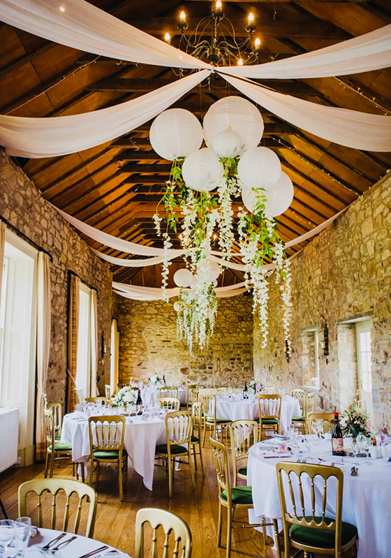 Barn wedding reception setup with round tables, draped fabric and hanging lanterns