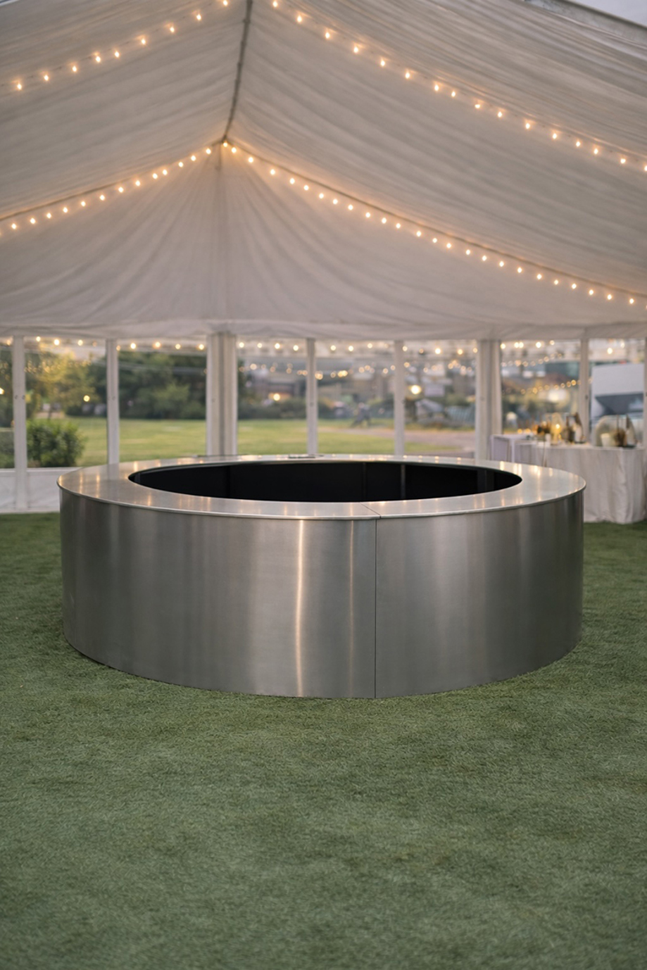 Circular stainless steel wedding bar by Sips Events styled inside a marquee with festoon lighting.