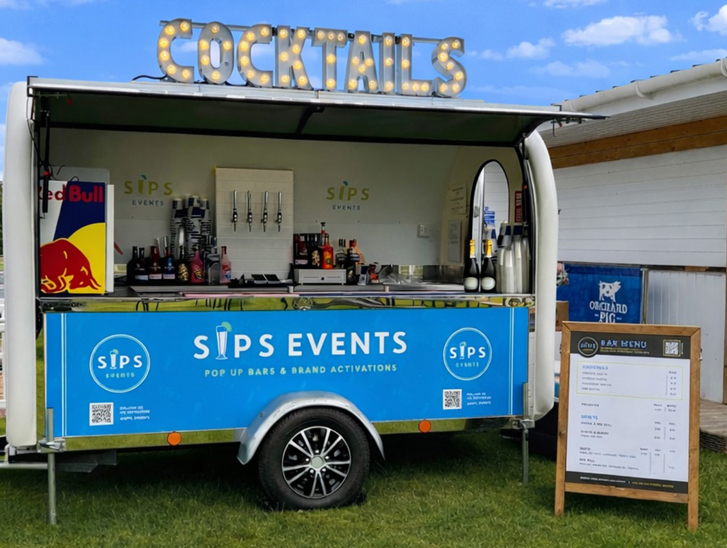 Mobile cocktail bar trailer by Sips Events set up outdoors with illuminated cocktail signage and drinks service station.