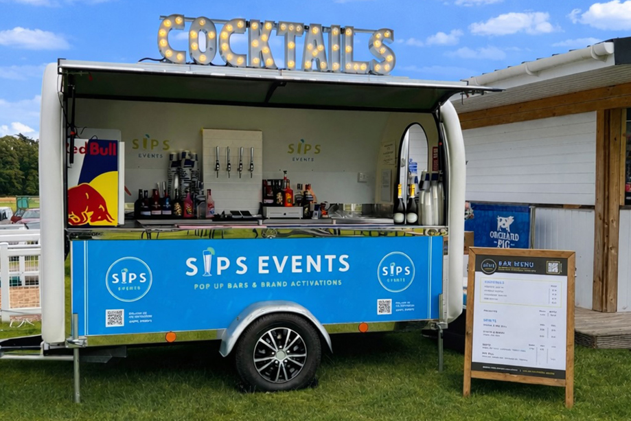 Mobile cocktail bar trailer by Sips Events set up outdoors with illuminated cocktail signage and drinks service station.