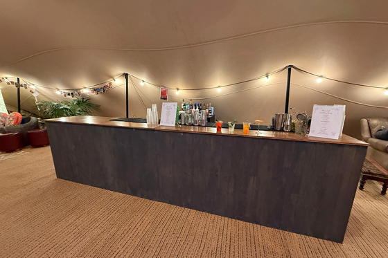 Rustic wooden drinks bar by Sips Events set up inside a tipi wedding venue with cocktails and menu displays.