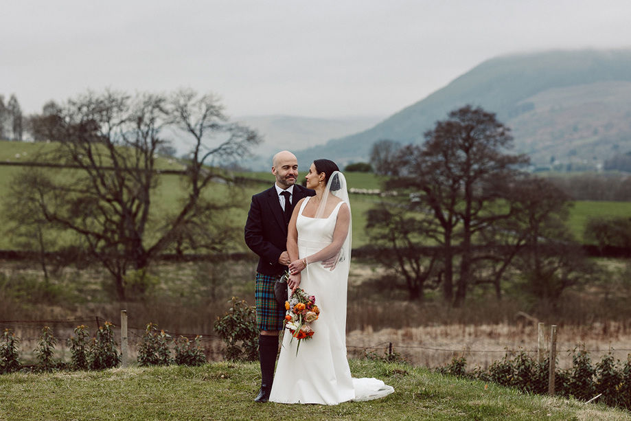 Bride and groom stand on the grass, looking into each others eyes with hills in background